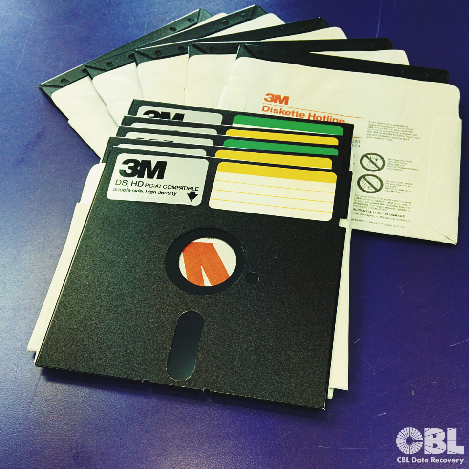 a stack of 5¼-inch floppy disks spread out on a purple counter. Their black plastic and white paper sleeves visible. Each disk has a yellow or green writeable label stickers in its top right corner and on the left corner a silver label with black text and logo: 3M, DS, HD PC/AT COMPATIBLE. double side, high density. CBL Data Recovery logo in bottom right corner of the image.