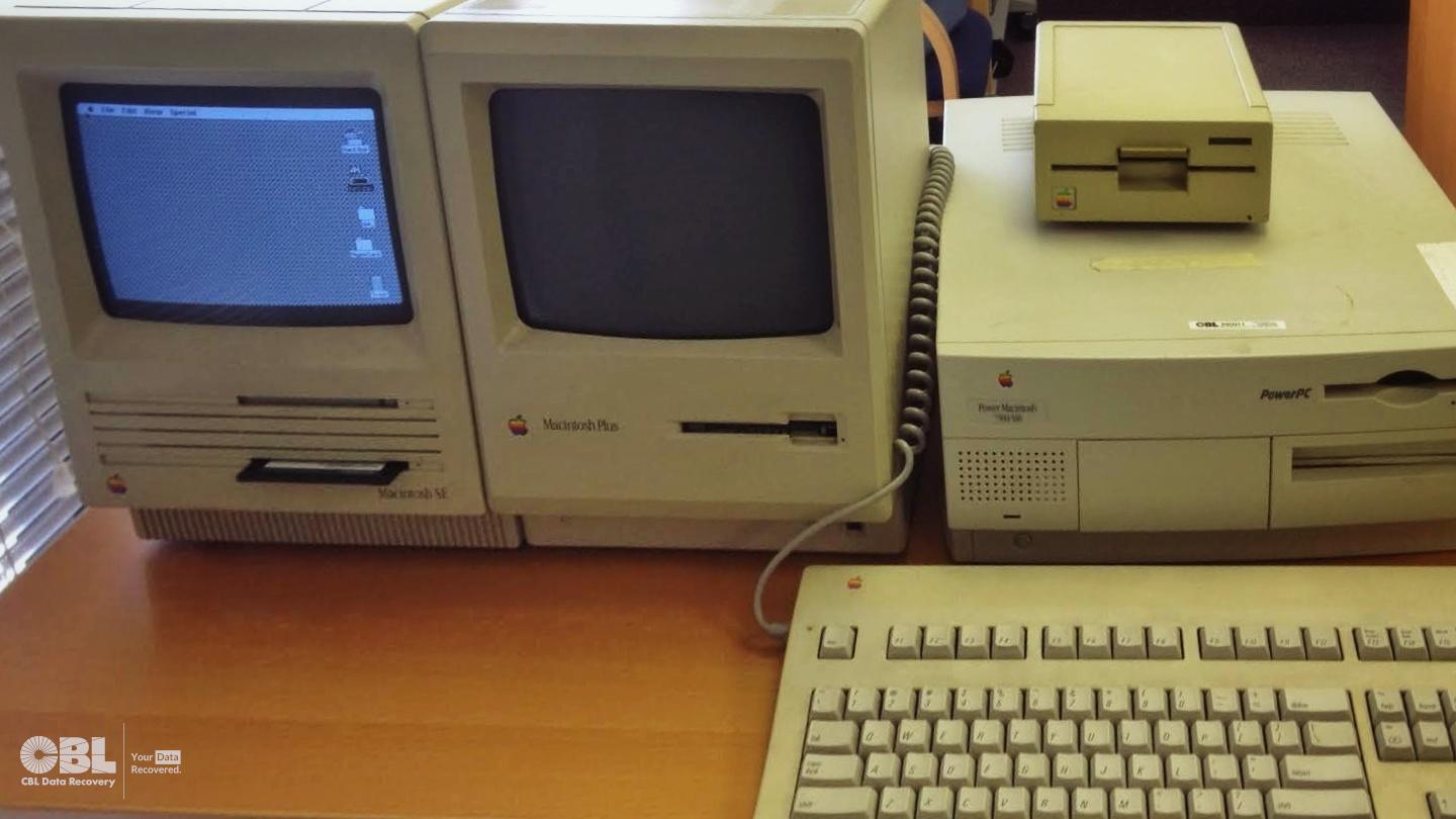 Some very old Apple Mac computers seen at CBL Data Recovery lab booted up on a table for a data recovery project. Macintosh SE, Macintosh Plus, and a Power Mac with 5.25 inch floppy drive external. Their cases all have a greyish-yellow tinge due to age! Old Mac OS version displayed on screen with very retro looking desktop. Distinct 'rainbow' Apple logos visible on each computer too.